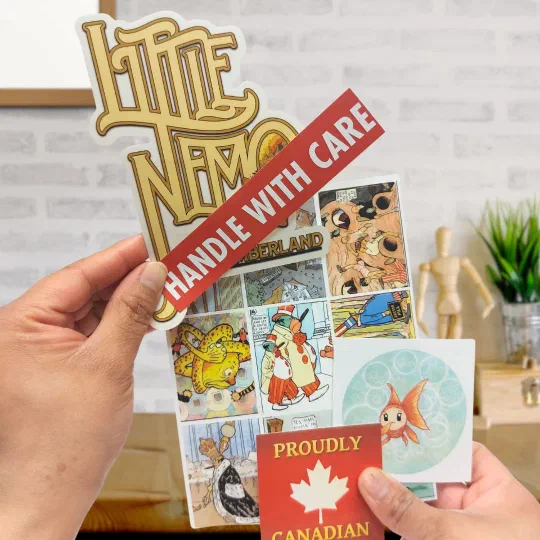 Die-cut stickers held in two hands: a Little Nemo wordmark, a “Handle with care” strip and sheets of comic panels